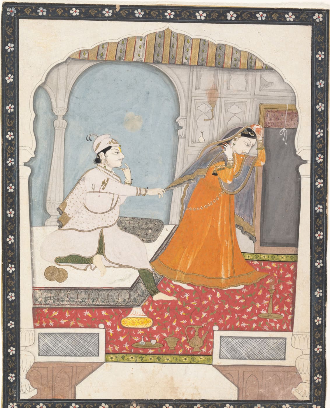 A young groom tugs at the orange chunni of his shy bride in a palace room